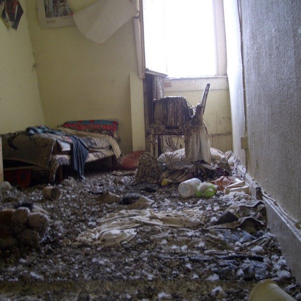 Blick in ein Schlafzimmer mit vereinzelten Möbelstücken, einem kleinen Fernseher und geöffneten Fenster. Der Boden ist massiv von Taubenkot durchtränkt.